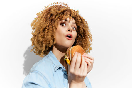 Happy woman with curly hair excitedly holding a burger, wearing a denim shirt against a white...