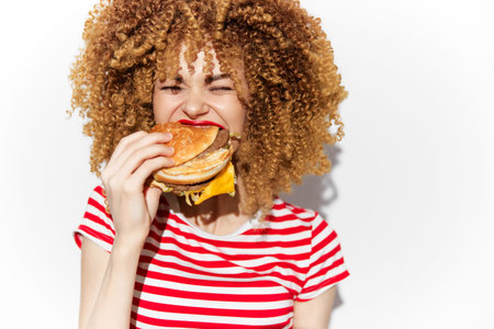Young woman with curly hair enjoying a cheesy burger, wearing a striped shirt, conveying happiness...