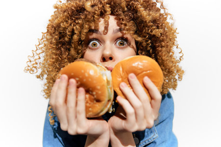 Young woman with curly hair excitedly holding two delicious burgers against a white background, showcasing vibrant colors and a playful moodの写真素材