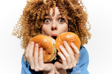 Surprised young woman with curly hair holding two burgers, showcasing excitement and joy in a...