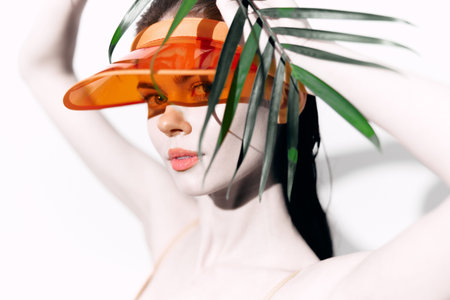Young woman in orange visor with palm leaves, exuding confidence and summer vibes in a bright, minimalist studio setting.の写真素材