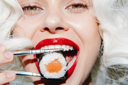 Woman with blonde curly hair enjoying sushi with chopsticks, showcasing vibrant lips and expressive eyes.の写真素材
