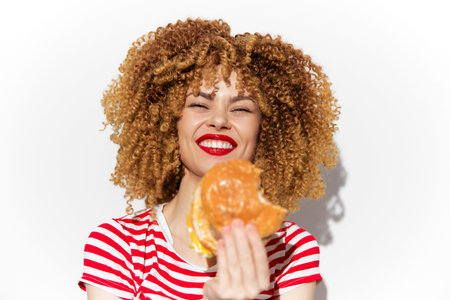 Happy young woman with curly hair enjoying a delicious burger against a light background, showcasing...