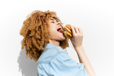Young woman enjoying a delicious burger with curly hair, showcasing a sense of happiness and indulgence against a clean white background Perfect for food and lifestyle themesの写真素材