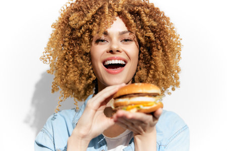Cheerful young woman with curly hair holding a delicious burger, emphasizing joy in indulgent eating, vibrant colors on a plain backgroundの写真素材