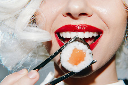 Glamorous woman enjoying sushi with red lips and white hair, showcasing exquisite sushi on chopsticks, vibrant colors, and joyful expression.の写真素材