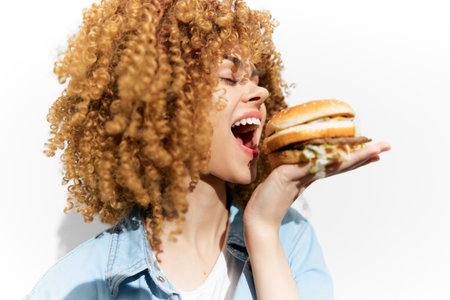 Young woman with curly hair joyfully holding a burger, expressive emotion, bright colors, healthy eating concept, seasonal appeal in a clean backgroundの写真素材