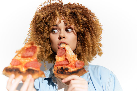 Young woman with curly hair enjoying pepperoni pizza, showcasing excitement in a bright, cheerful setting Perfect for food and lifestyle themesの写真素材