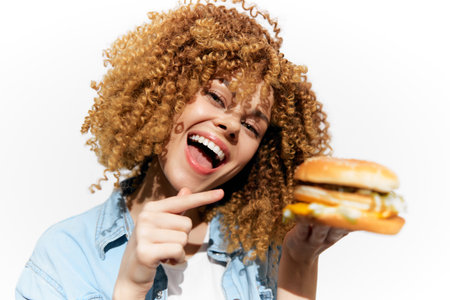 Cheerful young woman with curly hair holding a delicious hamburger against a bright white background, showcasing joy and enthusiasm for eatingの写真素材