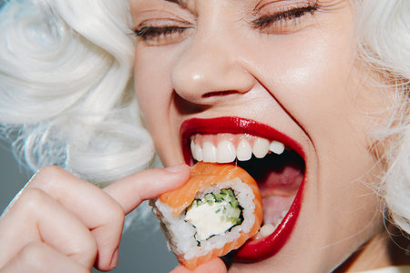 Smiling woman with curly hair enjoying sushi, vibrant red lips and playful expression, colorful and joyful image of culinary delight.の写真素材