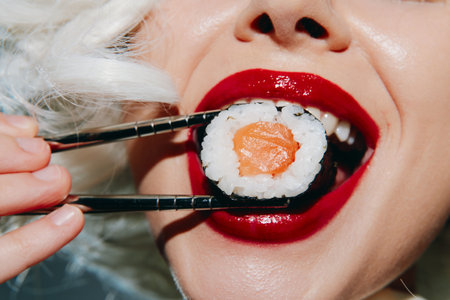 Brightly colored sushi being held by a woman with bold red lipstick, embodying enjoyment and culinary delight in a vibrant setting.の写真素材