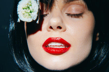 Close-up of a woman with vibrant red lips and sushi, expressing joy and style in a dark, modern setting, showcasing a unique culinary concept.の写真素材