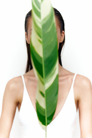 Fashion model obscured by a large green leaf, showcasing minimalist style and natural beauty on a white background, embodying tranquility and elegance.の写真素材