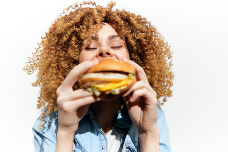 Happy young woman with curly hair enjoys a delicious hamburger against a bright white background, showcasing the joy of eating and indulgenceの写真素材