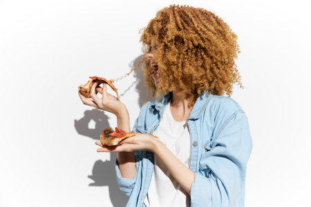 Happy young woman enjoying delicious pizza slices with curly hair, wearing a casual denim jacket against a white background, evoking a fun and carefree moodの写真素材