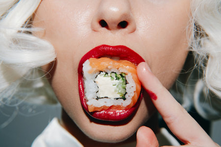 Close-up portrait of a woman with bold red lips holding sushi, showcasing vibrant colors and playful emotions for food enthusiasts.の写真素材