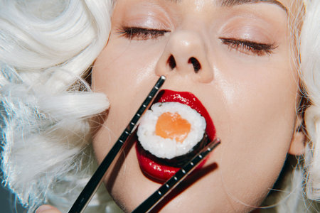 Glamorous close-up of a woman enjoying sushi with vibrant red lips and soft curly hair, showcasing elegance and indulgence in culinary delights.の写真素材