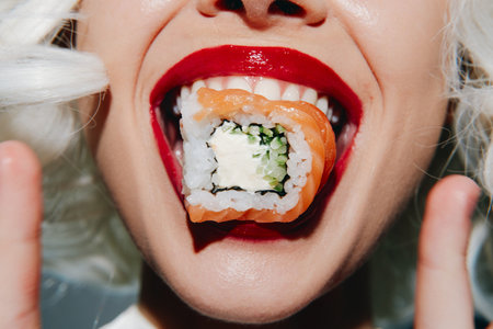 Playful woman with vibrant red lips playfully holding sushi in her mouth, showcasing joy and...