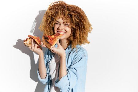 Cheerful young woman with curly hair holding two slices of pizza, embodying the joy of eating and indulgence in comfort foodの写真素材