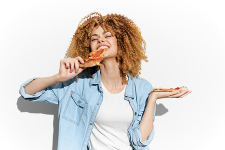 Joyful young woman enjoying pizza against a bright background, wearing a denim jacket, showcasing happiness and indulgence in a casual settingの写真素材