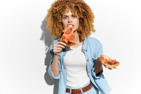 Young woman with curly hair enjoying pizza, showcasing emotions of indulgence and happiness, dressed...