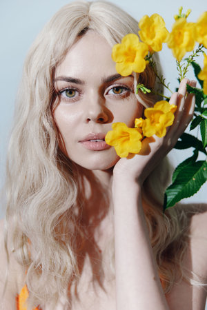 Young woman with blonde hair and green eyes posing with yellow flowers, wearing an orange outfit against a soft blue background.の写真素材
