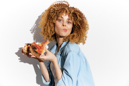 Happy young woman with curly hair holding a slice of pizza against a white background, showcasing a fun and vibrant lifestyle with a touch of indulgenceの写真素材