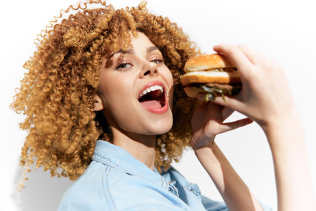 Joyful young woman with curly hair eating a delicious burger against a white background, showcasing a vibrant lifestyle and love for fast foodの写真素材