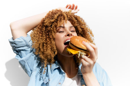 Happy young woman with curly hair enjoying a burger, bright and colorful, expressing joy in a casual style. Ideal for food-related content and lifestyle imagery.の写真素材