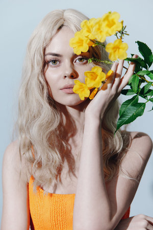 Beautiful young woman with long blonde hair holding yellow flowers against a light blue background in a bright orange dress, conveying joy and freshness.の写真素材