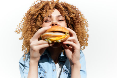 Young woman with curly hair enjoying a delicious burger in a bright setting, capturing the joy of eating and indulgence with a vibrant atmosphereの写真素材
