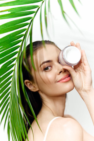 Beauty portrait of a model holding a skincare cream with palm leaves, exuding freshness and natural beauty in a bright, tropical setting.の写真素材