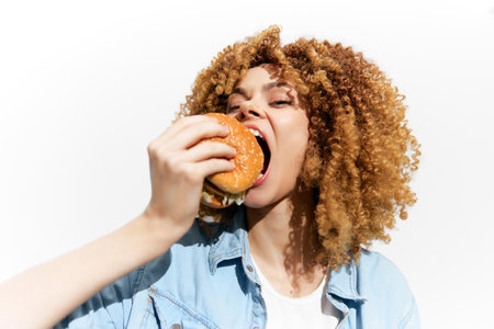 Joyful woman with curly hair savoring a delicious burger against a bright white background, embodying a carefree attitude towards eating and indulgenceの写真素材