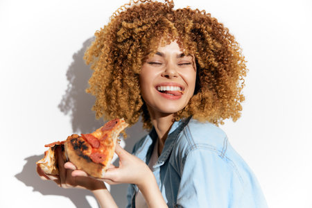 Happy young woman with curly hair enjoying pizza, wearing a denim shirt, bright smile, isolated on white background, perfect for food and lifestyle conceptsの写真素材