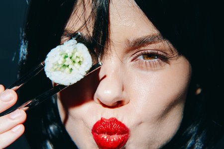 Young woman with dark hair holding sushi roll, expressing joy and vitality with vibrant red lipstick...