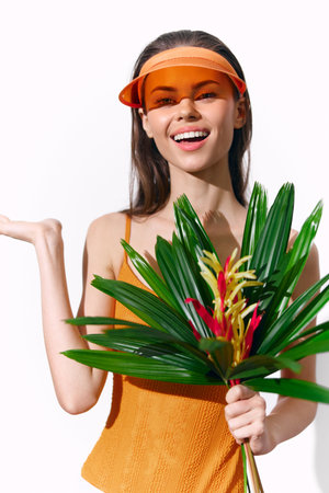 Cheerful young woman in orange outfit with visor holds tropical leaves and flowers against a bright background, perfect for summer concepts.の写真素材