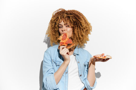 Young woman enjoying pizza with curly hair, wearing a denim jacket, showcasing happiness against a white background, ideal for food and lifestyle themesの写真素材