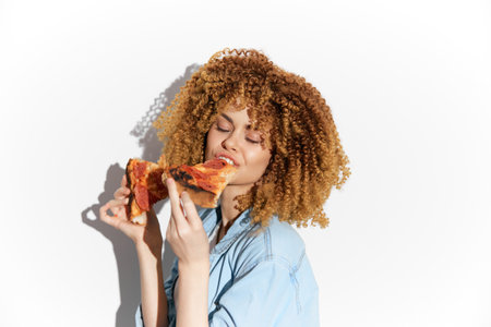 Young woman enjoying a slice of pizza with curly hair, dressed in denim shirt, expressing joy against a white background, perfect for food related conceptsの写真素材