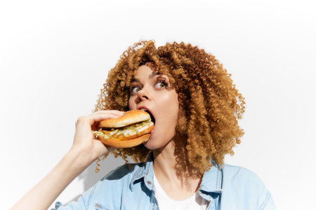 Young woman with curly hair excitedly eating a delicious burger, showcasing a vibrant and playful attitude, perfect for fast food imageryの写真素材