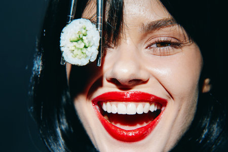 Joyful woman holding sushi with chopsticks, showcasing vibrant red lips and a cheerful expression against a dark background. Perfect for food and lifestyle concepts.の写真素材