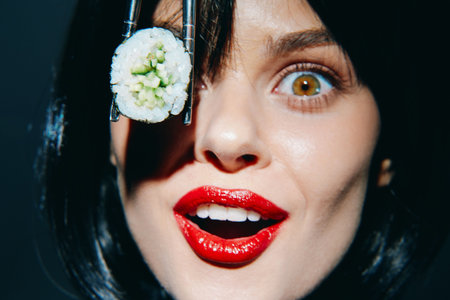 Excited woman holding sushi with chopsticks, showcasing vibrant emotion and colorful makeup against a dark background.の写真素材