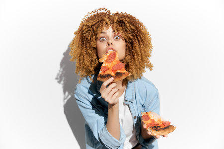 Young woman with curly hair joyfully eating pizza, showcasing excitement and love for food against a plain backgroundの写真素材
