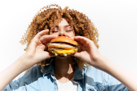 Young woman with curly hair joyfully holding a delicious burger, showcasing a vibrant and energetic expression, perfect for fast food promotionsの写真素材
