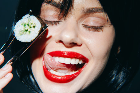 Playful young model enjoying sushi with vibrant red lips and a joyful expression against a dark...