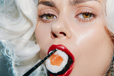 Stunning woman with sushi: close-up portrait highlighting vibrant sushi roll and bold red lips, showcasing food art and modern culinary trends.の写真素材
