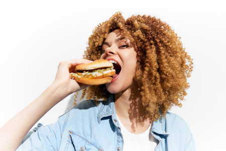 Young woman with curly hair enjoying a delicious burger, showcasing joy and satisfaction in a bright, minimalistic background Fast food, lifestyle, summer vibeの写真素材