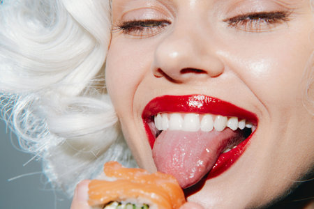 Playful woman enjoying sushi with a teasing smile, showcasing vibrant red lips and a cheeky expression against a soft background.の写真素材
