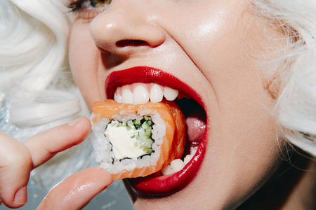 Model with blonde curls enjoying sushi, bright red lips, and joyful expression, set against a soft background. A colorful culinary delight capturing a moment of indulgence.の写真素材