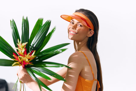Young woman in bright orange swimwear holding tropical leaves and flowers, smiling joyfully against a white background. Perfect for summer vibes and tropical themes.の写真素材