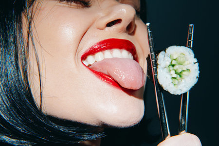 Playful woman teasingly tasting sushi with chopsticks, showcasing vibrant red lips and a confident expression against a dark background.の写真素材